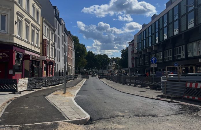 Bild Eppendorfer Marktplatz Das Bild zeigt eine Straße am Eppendorfer Marktplatz. Sie ist verschmutzt von Bauarbeiten. Links und rechts der Straße sind Wohnhäuser und Geschäfte zu sehen. Der Himmel ist blau. Die Sonne scheint.