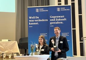 Zwei Personen stehen vor großen blauen Bannern der HAW und halten Blumentöpfe in der Hand.